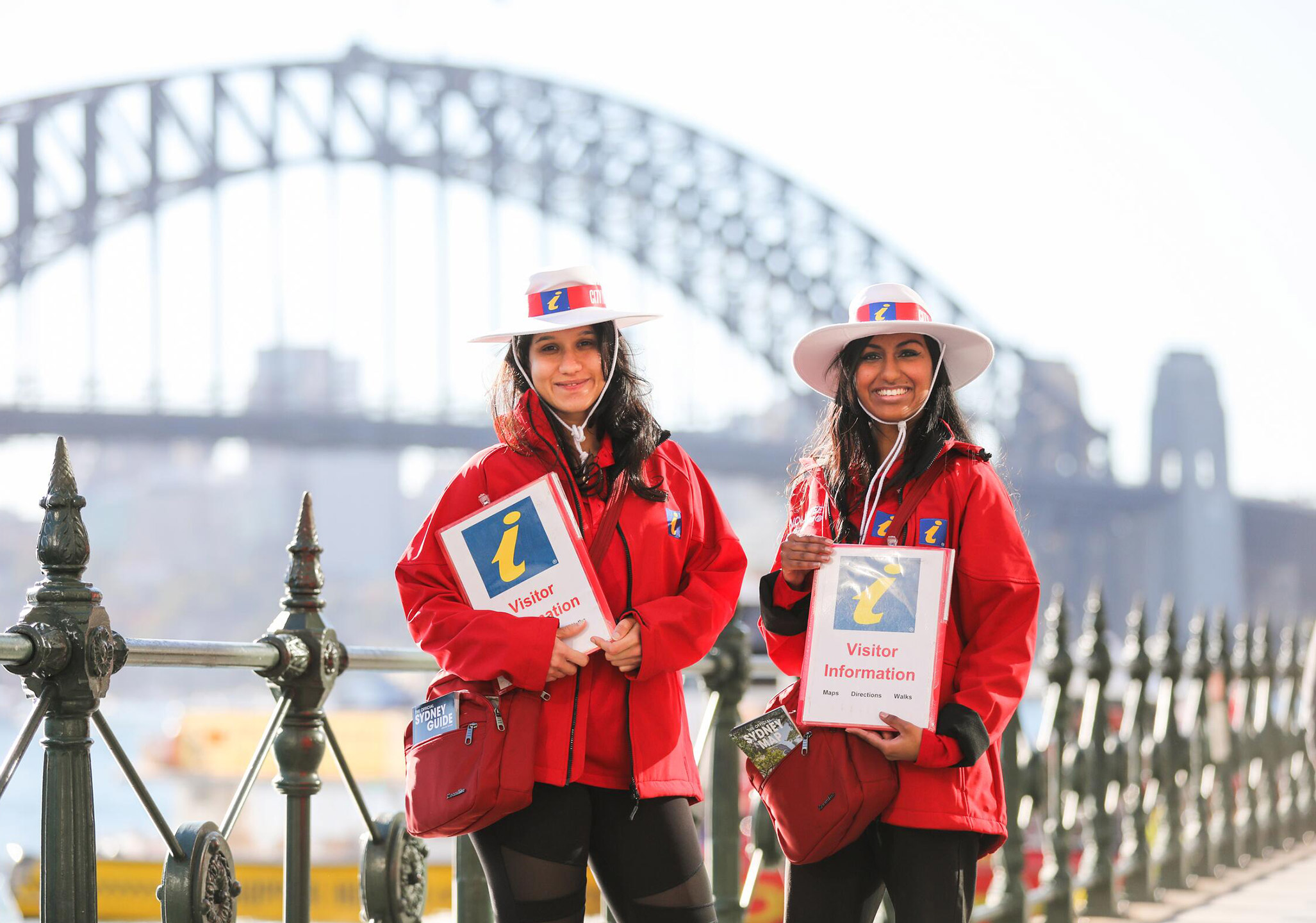 Two women in red uniforms and hats hold visitor information signs near the Sydney Harbour Bridge on a sunny day.